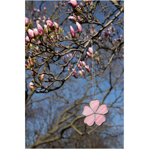 Handheld handcrafted Sakura cherry blossom suncatcher made of pink and yellow opalescent stained glass against a blurry spring tree background.