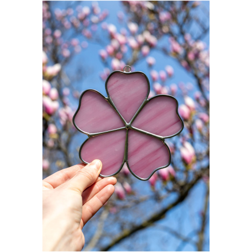 Handheld handcrafted Sakura cherry blossom suncatcher made of pink and yellow opalescent stained glass against a blurry spring tree background.