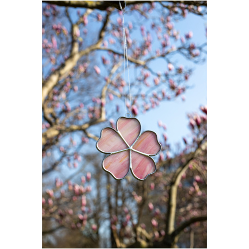 Handheld handcrafted Sakura cherry blossom suncatcher made of pink and yellow opalescent stained glass against a blurry spring tree background.