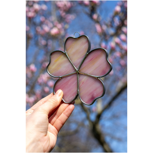 Handheld handcrafted Sakura cherry blossom suncatcher made of pink and yellow opalescent stained glass against a blurry spring tree background.